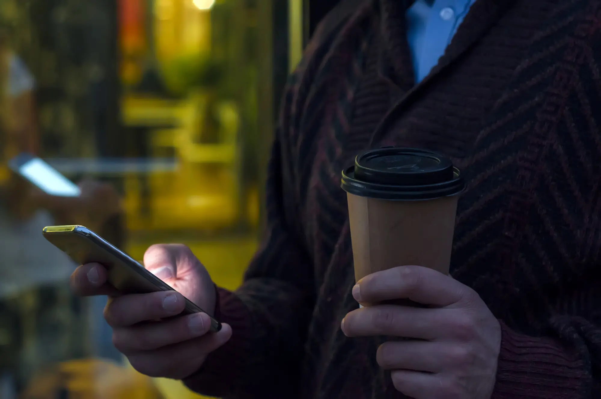 Nahaufnahme eines Geschäftsmanns, der ein Mobiltelefon benutzt und einen Pappbecher hält. Detailaufnahme der Hand des Geschäftsmanns, die einen Pappbecher hält und ein Smartphone benutzt, während er auf der Straße geht.